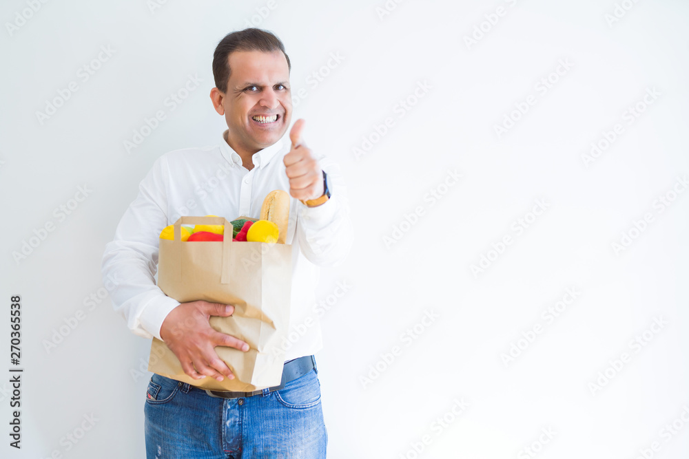 Middle age man holding groceries shopping bag over white background happy with big smile doing ok sign, thumb up with fingers, excellent sign Middle age man holding groceries shopping bag over white background happy with big smile doing ok sign, thumb up with fingers, excellent sign