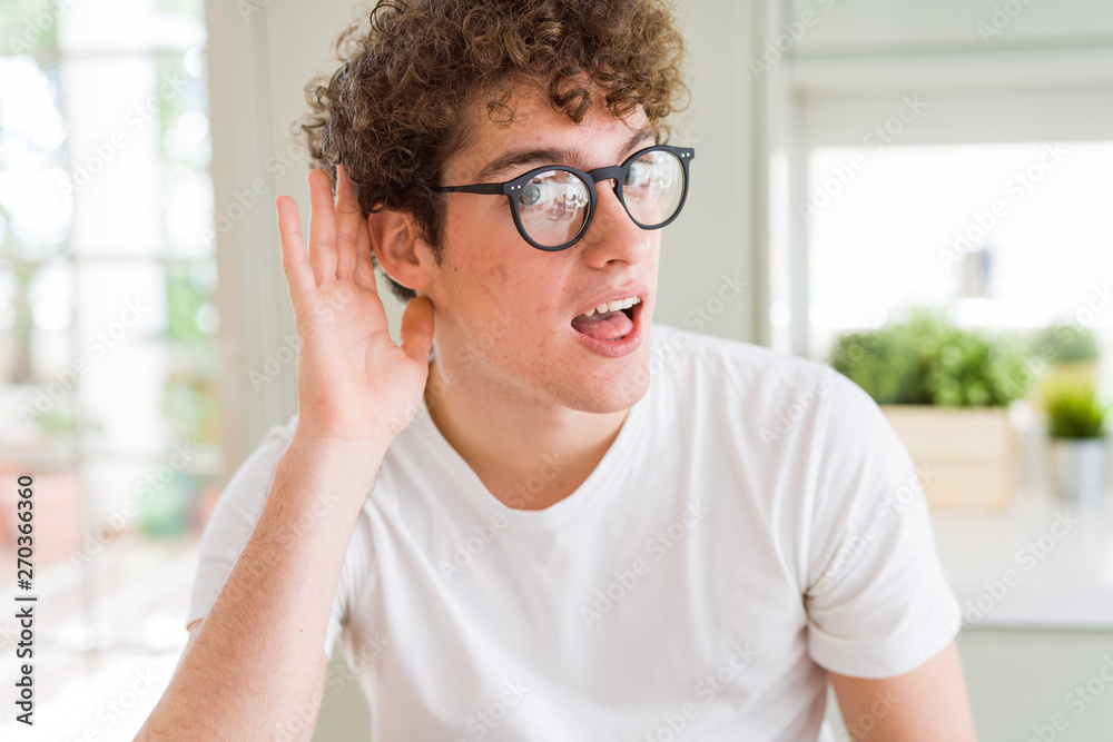 Young handsome man wearing glasses smiling with hand over ear listening an hearing to rumor or gossip. Deafness concept. Young handsome man wearing glasses smiling with hand over ear listening an hearing to rumor or gossip. Deafness concept.