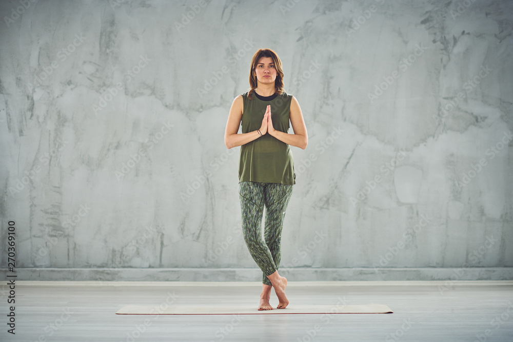 Full length of charming fit Caucasian brunette dressed in green outfit standing on mat and doing yoga. Full length of charming fit Caucasian brunette dressed in green outfit standing on mat and doing yoga.