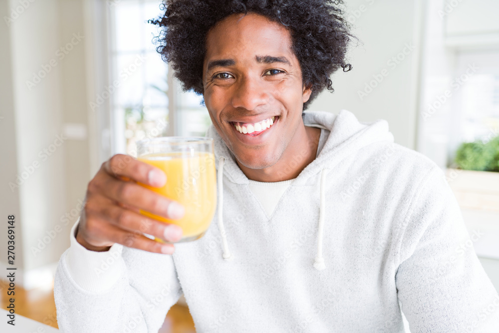 African American man holding and drinking glass of orange juice with a happy face standing and smiling with a confident smile showing teeth African American man holding and drinking glass of orange juice with a happy face standing and smiling with a confident smile showing teeth