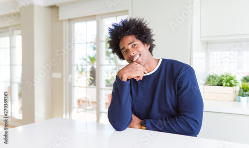 Young african american man wearing casual sweater sitting at home looking confident at the camera smiling with crossed arms and hand raised on chin. Thinking positive. © Krakenimages.com