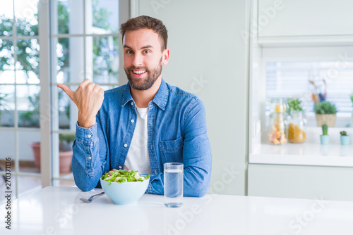 Handsome man eating fresh healthy salad smiling with happy face looking and pointing to the side with thumb up.