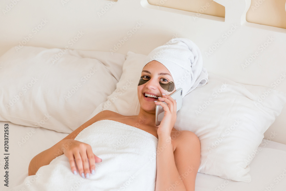 Beautiful smiling young woman with under eye patches and talking mobile phone in bathrobe lying in bed. Happy girl taking care of herself. Beauty skincare and wellness morning concept Beautiful smiling young woman with under eye patches and talking mobile phone in bathrobe lying in bed. Happy girl taking care of herself. Beauty skincare and wellness morning concept