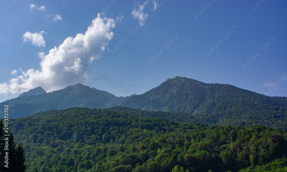 Summer in the mountains. The mountain slopes are covered with greenery. A cloud is emerging from behind the mountains.
