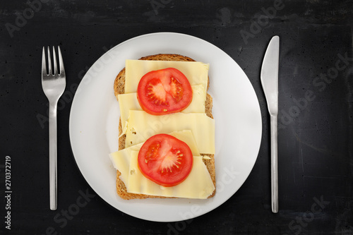 Slice of brown bread with cheese and tomato on white plate, stainless steel silverware and black stone background.