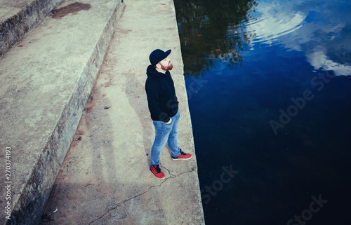 Hipster handsome male model with beard wearing black hoodie and cap in casual urban style 