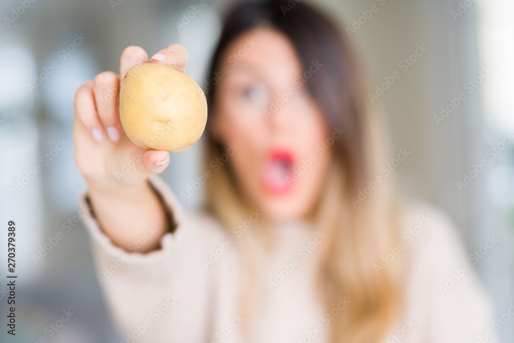 Young beautiful woman holding fresh potato at home scared in shock with a surprise face, afraid and excited with fear expression Young beautiful woman holding fresh potato at home scared in shock with a surprise face, afraid and excited with fear expression