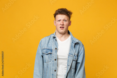 Portrait of shocked disgusted young man in denim casual clothes standing and looking camera isolated on yellow orange wall background. People sincere emotions, lifestyle concept. Mock up copy space.