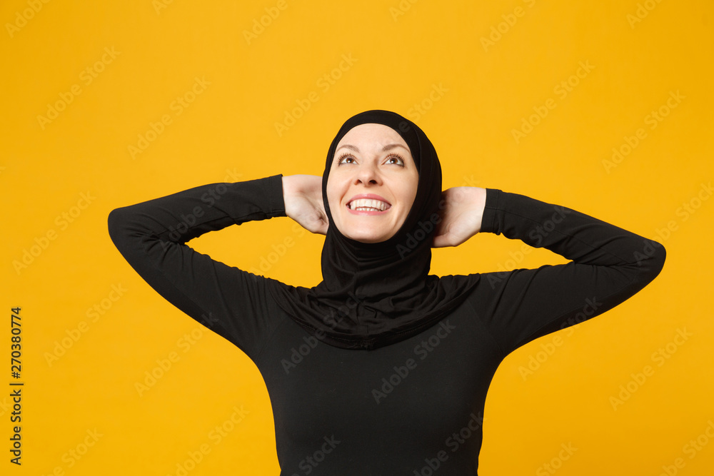 Smiling confident beautiful young arabian muslim woman in hijab black clothes posing isolated on yellow wall background, studio portrait. People religious Islam lifestyle concept. Mock up copy space.