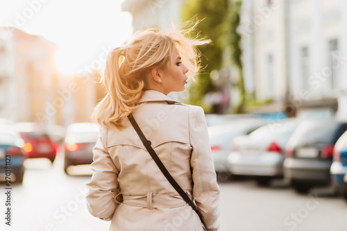 Outdoor portrait from back of serious lady in beige coat looking away standing on the street. Pretty girl with blonde ponytail walking around city in sunny day. © Look!