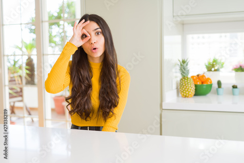 Young beautiful woman at home on white table doing ok gesture shocked with surprised face, eye looking through fingers. Unbelieving expression.