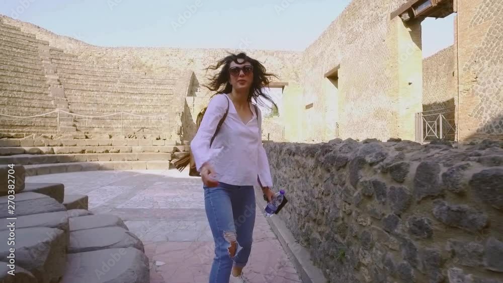 Back view of fit stylish young brunette woman walking down the ruins of ancient theatre, turns to camera and smiles happily. Enjoying the moment, happiness, positive. Follow me concept Back view of fit stylish young brunette woman walking down the ruins of ancient theatre, turns to camera and smiles happily. Enjoying the moment, happiness, positive. Follow me concept