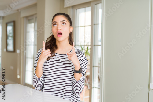Young beautiful woman at home amazed and surprised looking up and pointing with fingers and raised arms.