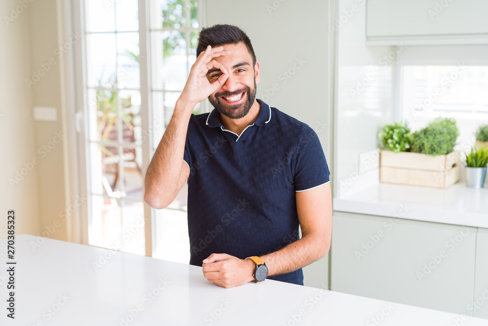 Handsome hispanic man at home doing ok gesture with hand smiling, eye looking through fingers with happy face.