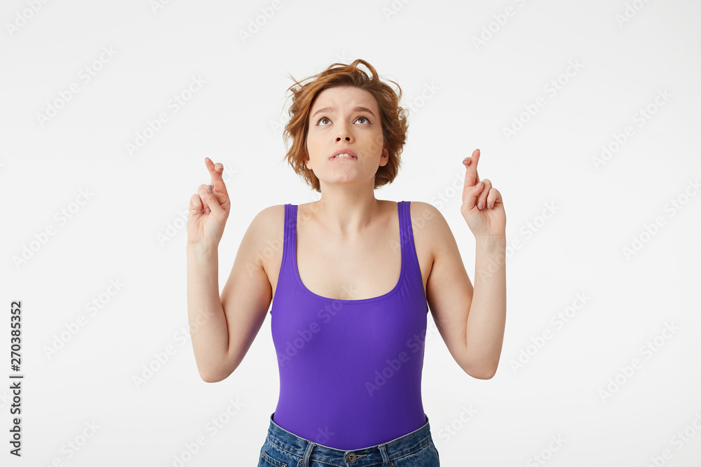 Photo of young nice short-haired girl, fingers crossed and lips pressed upwards and looks forward to good luck in the exam, stands over white wall. Photo of young nice short-haired girl, fingers crossed and lips pressed upwards and looks forward to good luck in the exam, stands over white wall.