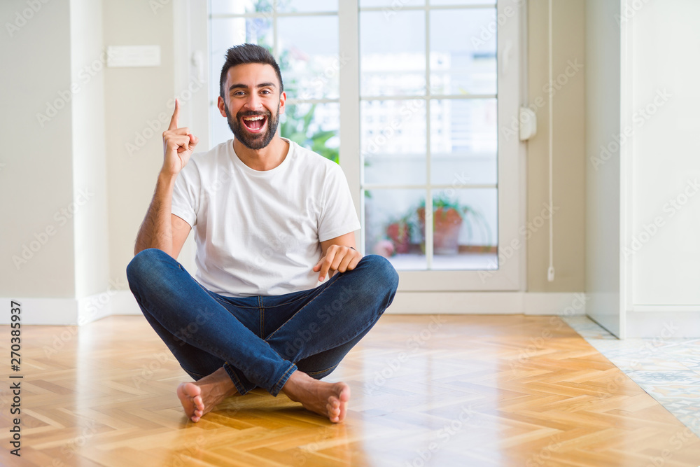 Handsome hispanic man wearing casual t-shirt sitting on the floor at home pointing finger up with successful idea. Exited and happy. Number one.