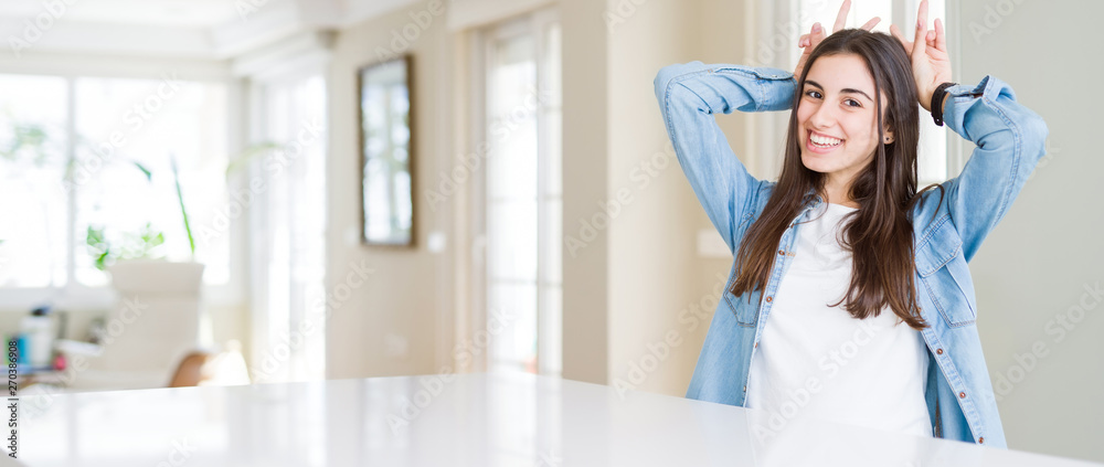 Wide angle picture of beautiful young woman sitting on white table at home Posing funny and crazy with fingers on head as bunny ears, smiling cheerful Wide angle picture of beautiful young woman sitting on white table at home Posing funny and crazy with fingers on head as bunny ears, smiling cheerful