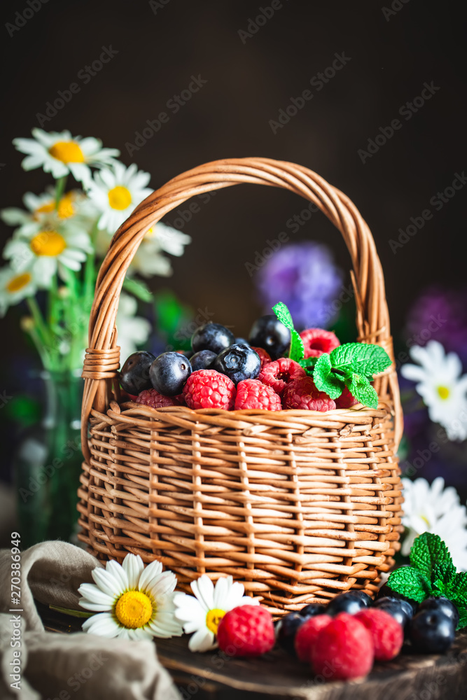 Raspberries and blueberries in a basket with chamomile and leaves on a dark background. Summer and healthy food concept. Selective focus. Background with copy space. Raspberries and blueberries in a basket with chamomile and leaves on a dark background. Summer and healthy food concept. Selective focus. Background with copy space.