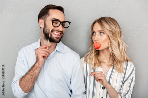 Photo closeup of nice couple in casual clothing looking at each other and holding paper fake glasses and lips on stick