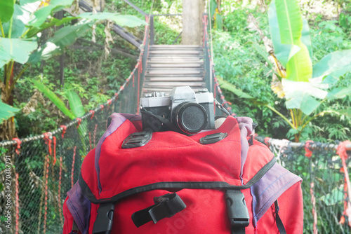 Camera lay on red backpack with rope bridge on background, adventure concept.