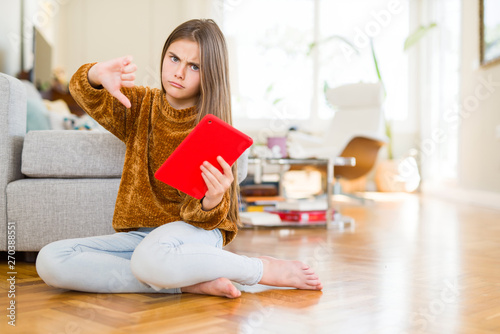 Beautiful young girl kid using digital touchpad tablet sitting on the floor with angry face, negative sign showing dislike with thumbs down, rejection concept