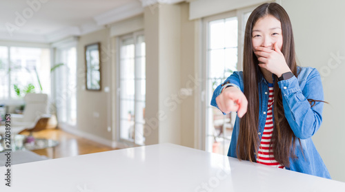 Young beautiful asian woman with long hair wearing denim jacket laughing at you  pointing finger to the camera with hand over mouth  shame expression