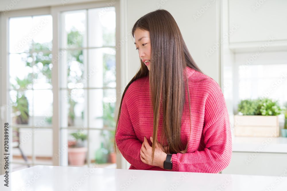 Beautiful Asian woman wearing pink sweater on white table with hand on stomach because nausea, painful disease feeling unwell. Ache concept.