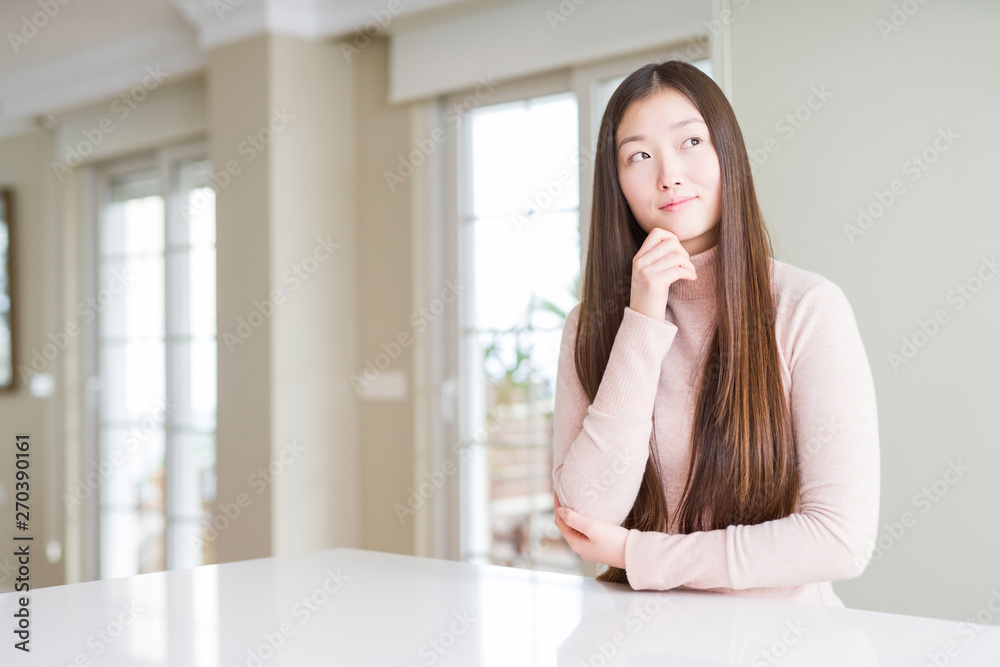 Beautiful Asian woman wearing casual sweater on white table with hand on chin thinking about question, pensive expression. Smiling with thoughtful face. Doubt concept. Beautiful Asian woman wearing casual sweater on white table with hand on chin thinking about question, pensive expression. Smiling with thoughtful face. Doubt concept.