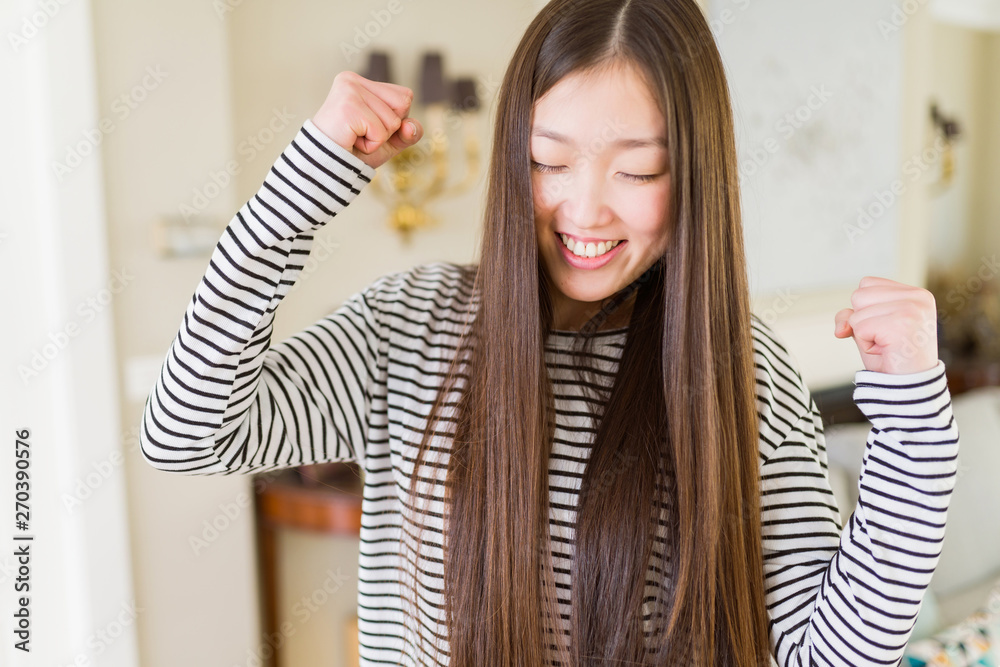 Beautiful Asian woman wearing stripes sweater over living room background excited for success with arms raised celebrating victory smiling. Winner concept. Beautiful Asian woman wearing stripes sweater over living room background excited for success with arms raised celebrating victory smiling. Winner concept.