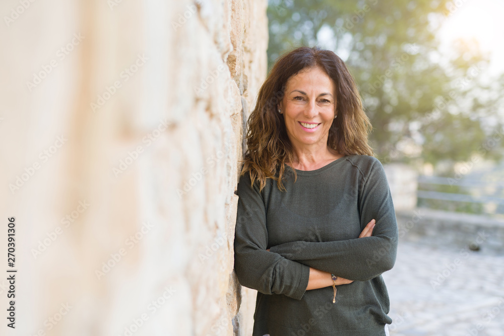 Beautiful middle age woman smiling cheerful leaning on a brick wall at the city street on a sunny day Beautiful middle age woman smiling cheerful leaning on a brick wall at the city street on a sunny day
