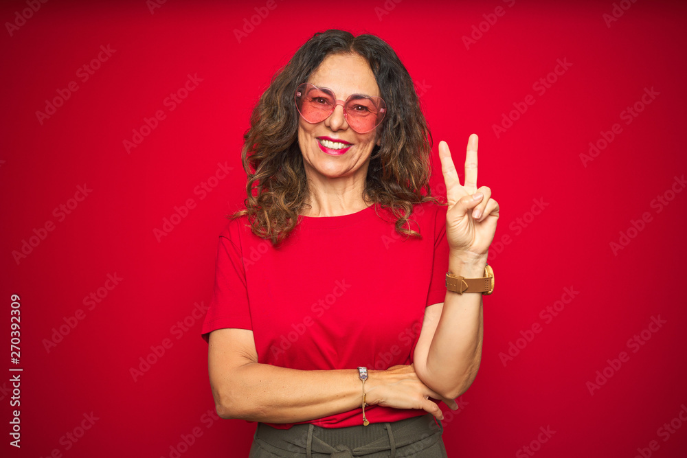 Middle age senior woman wearing cute heart shaped glasses over red isolated background smiling with happy face winking at the camera doing victory sign. Number two. Middle age senior woman wearing cute heart shaped glasses over red isolated background smiling with happy face winking at the camera doing victory sign. Number two.