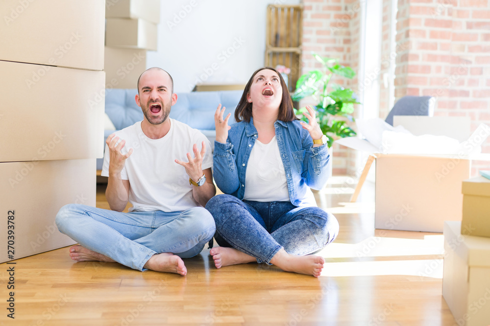Young couple sitting on the floor arround cardboard boxes moving to a new house crazy and mad shouting and yelling with aggressive expression and arms raised. Frustration concept.