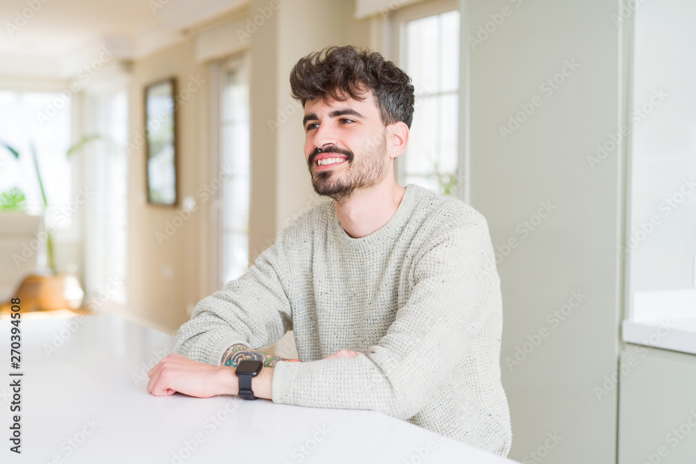 Young man wearing casual sweater sitting on white table looking away to side with smile on face, natural expression. Laughing confident.