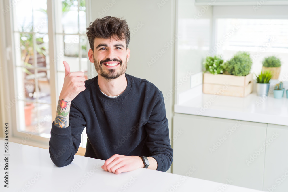 Young man wearing casual sweatshirt sitting on white table doing happy thumbs up gesture with hand. Approving expression looking at the camera with showing success. Young man wearing casual sweatshirt sitting on white table doing happy thumbs up gesture with hand. Approving expression looking at the camera with showing success.