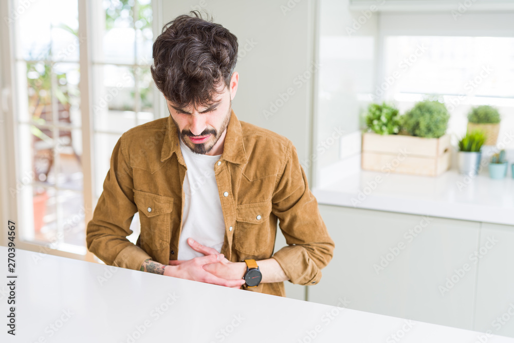Young man wearing casual jacket sitting on white table with hand on stomach because indigestion, painful illness feeling unwell. Ache concept.