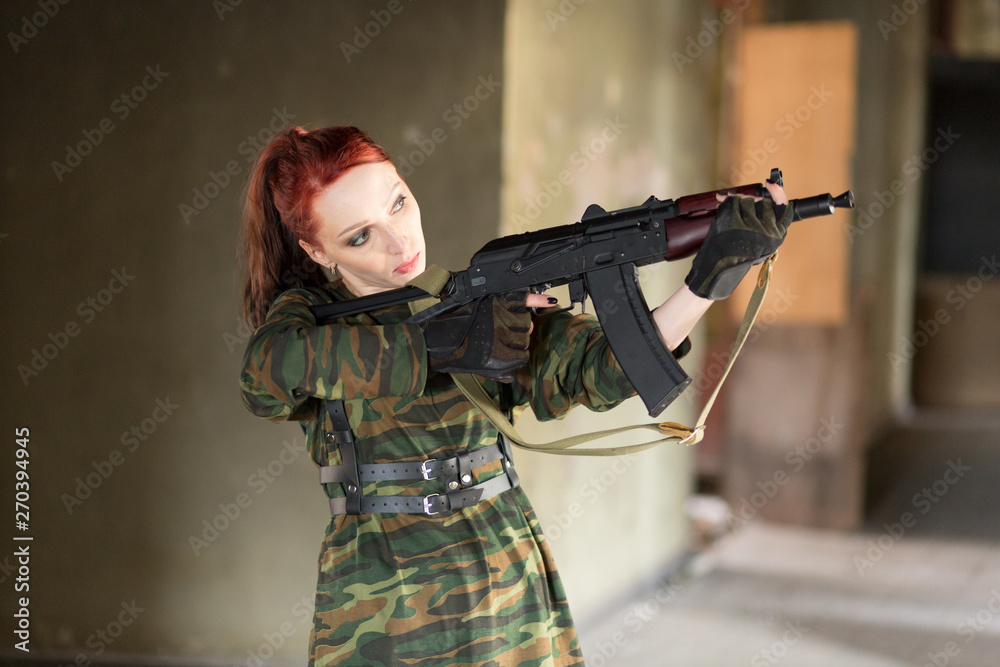 Fototapeta premium Girl with red hair with a gun standing in an abandoned building and looks away. A young girl in a camouflage t-shirt and a waistcoat. Girl in leather belts.