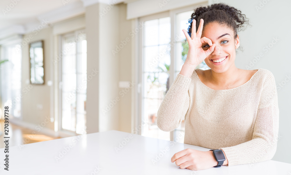 Beautiful young african american woman with afro hair sitting on table at home doing ok gesture with hand smiling, eye looking through fingers with happy face. Beautiful young african american woman with afro hair sitting on table at home doing ok gesture with hand smiling, eye looking through fingers with happy face.