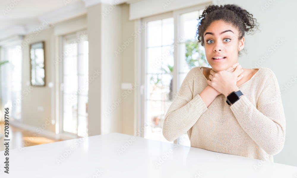 Beautiful young african american woman with afro hair sitting on table at home shouting and suffocate because painful strangle. Health problem. Asphyxiate and suicide concept.