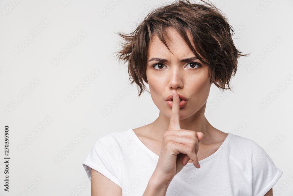 Portrait of strict woman with short brown hair in basic t-shirt holding index finger on lips Portrait of strict woman with short brown hair in basic t-shirt holding index finger on lips