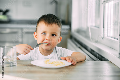 a little boy eats pasta in the form of a spiral in the afternoon in the kitchen on their own