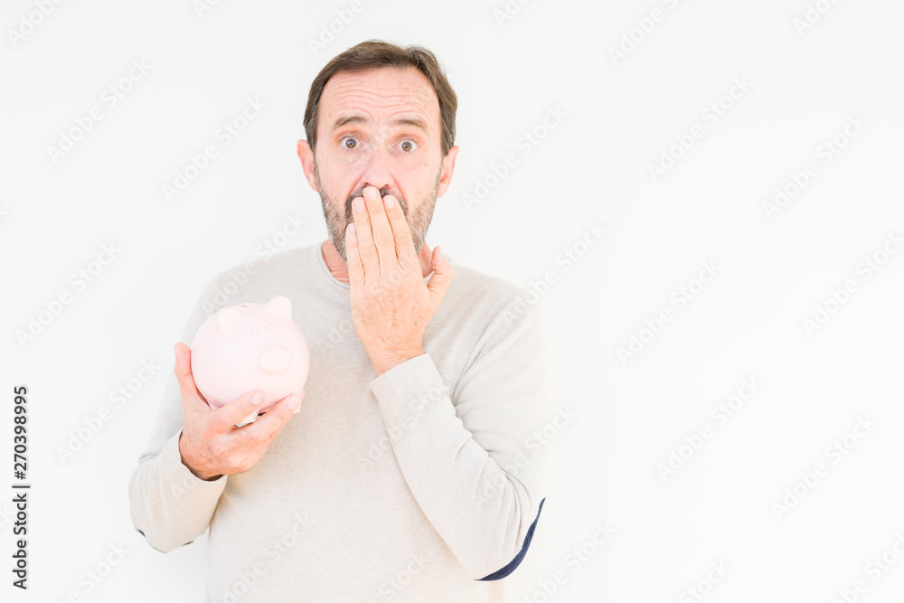 Senior man holding piggy bank over isolated background cover mouth with hand shocked with shame for mistake, expression of fear, scared in silence, secret concept Senior man holding piggy bank over isolated background cover mouth with hand shocked with shame for mistake, expression of fear, scared in silence, secret concept