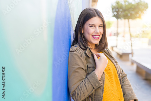 Beautiful young woman smiling confident and cheerful leaning on blue wall, walking on the street of the city on a sunny day