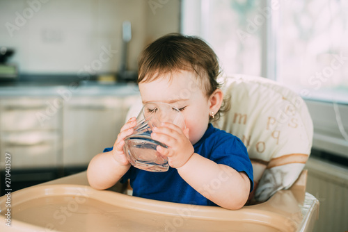 A little girl in the kitchen in the afternoon in a high chair drinking water from a glass glass