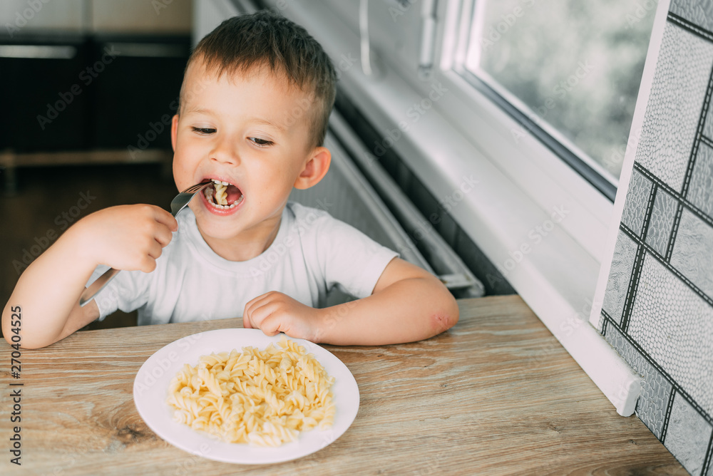 little boy eats pasta in the form of a spiral in the afternoon in the kitchen on their own little boy eats pasta in the form of a spiral in the afternoon in the kitchen on their own