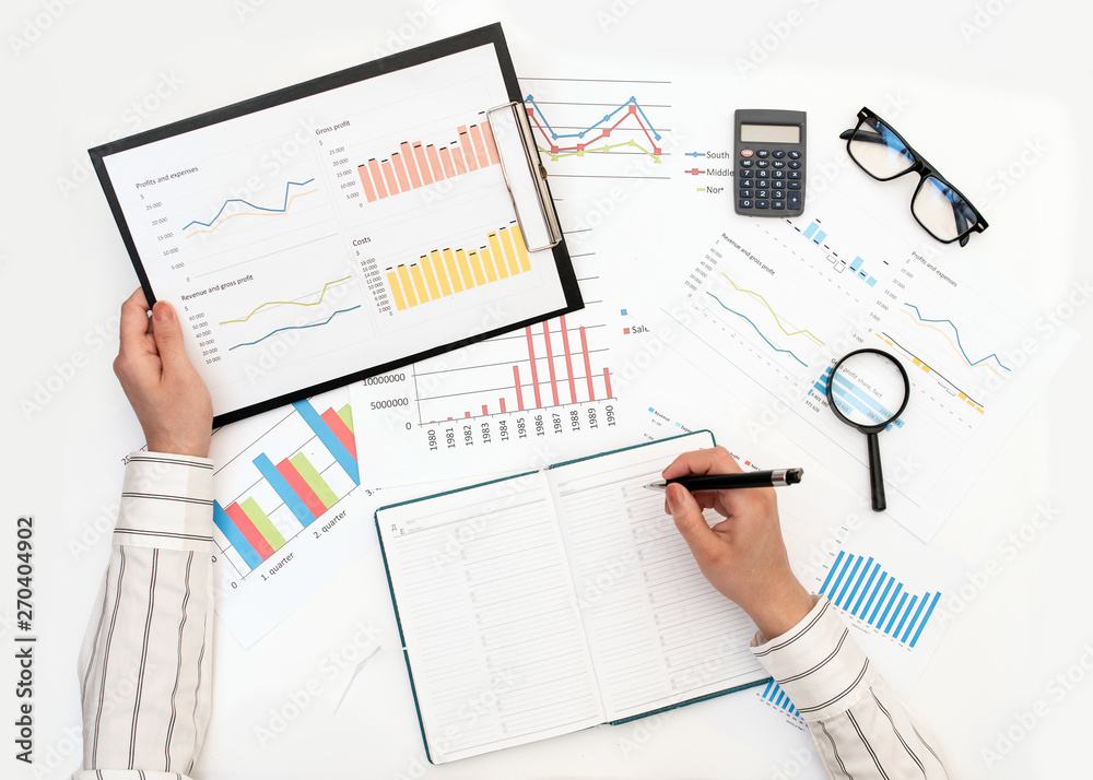 A woman's hands record data from a report with graphs. Top view on the hands on the white table. Finance and business concept. A woman's hands record data from a report with graphs. Top view on the hands on the white table. Finance and business concept.
