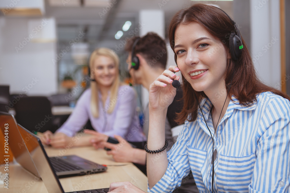 Cheerful helpful female call center operator smiling to the camera, working at the office, copy space. Charming young woman working at customer support service. Happy female at technical support offic