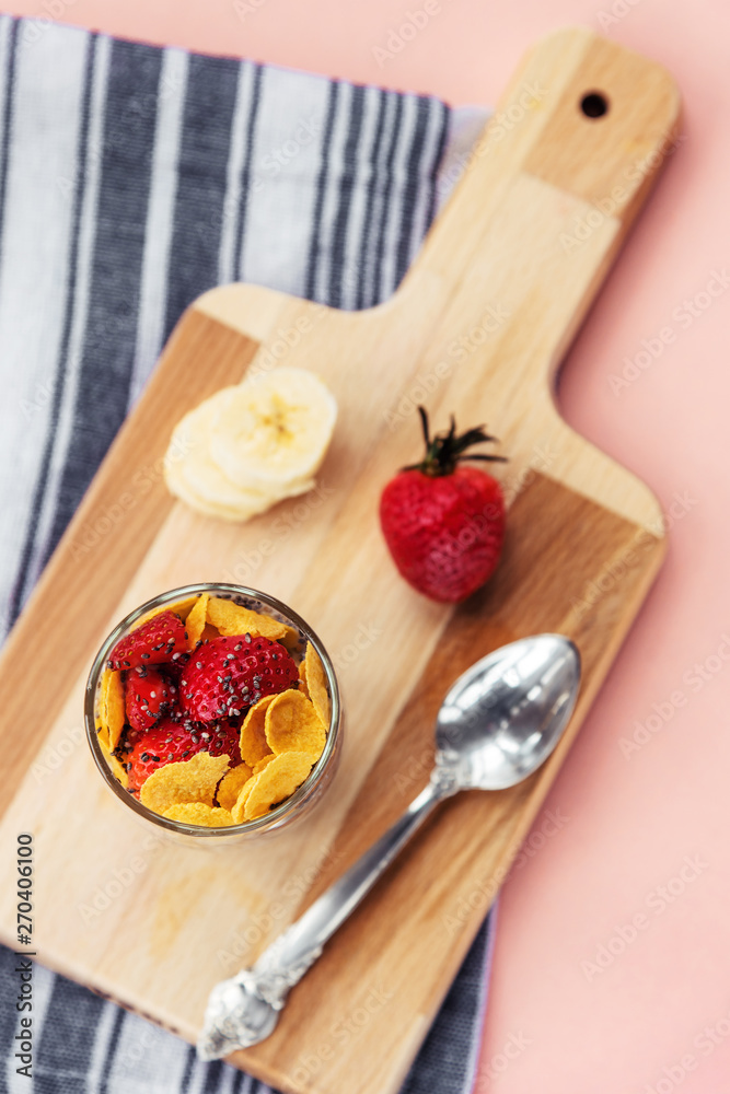 Top view of Cornflakes with banana, strawberry and chia seeds on a wooden cutting board on pink background, healthy breakfast. Soft focus. Top view of Cornflakes with banana, strawberry and chia seeds on a wooden cutting board on pink background, healthy breakfast. Soft focus.