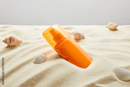 sunscreen in orange bottle in sand with seashells on grey background