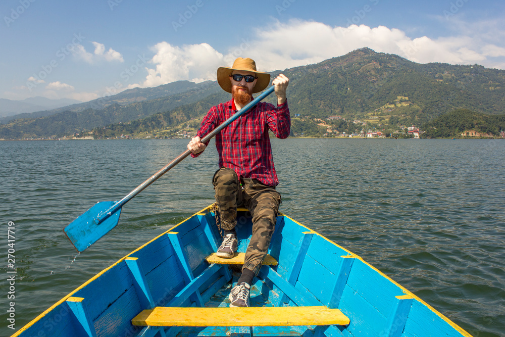 bearded man in a red shirt, hat and sunglasses in a blue wooden boat with a paddle in his hands, on the lake against the background of green mountains