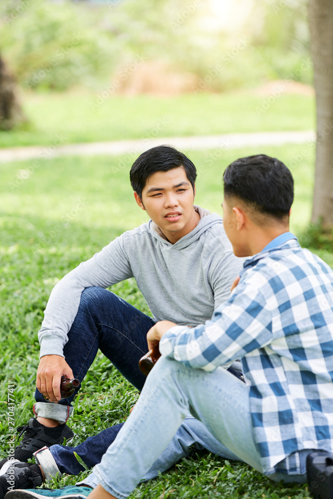 Two handsome young men drinking beer and talking to each other while sitting on the grass together Two handsome young men drinking beer and talking to each other while sitting on the grass together